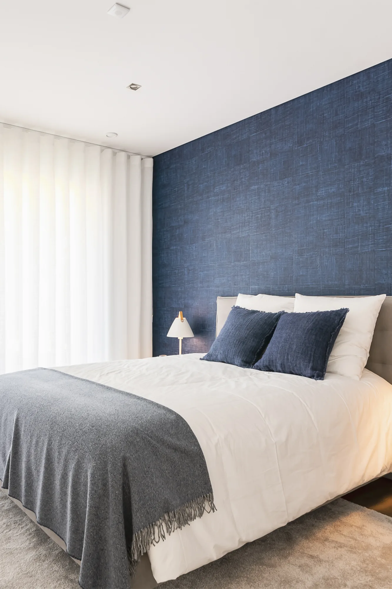 Quarto com parede de fundo em azul profundo, roupa de cama clara e cortinados.